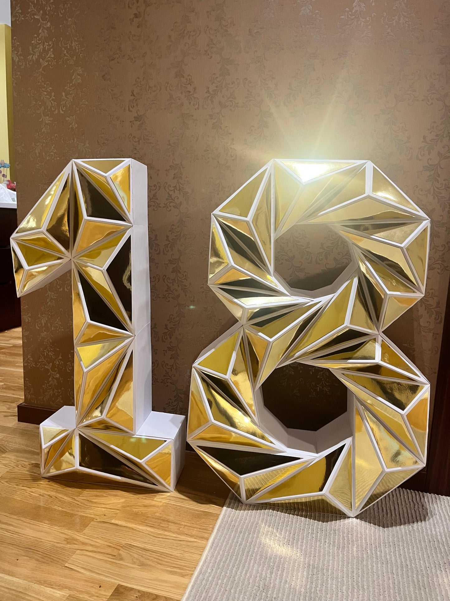 Ručno rađeni low poly brojevi 1 i 8 u zlatno-beloj kombinaciji, geometrijski dizajn sa trouglovima, idealni za 18. rođendan ili jubilej, postavljeni na drvenom stolu sa neutralnom pozadinom