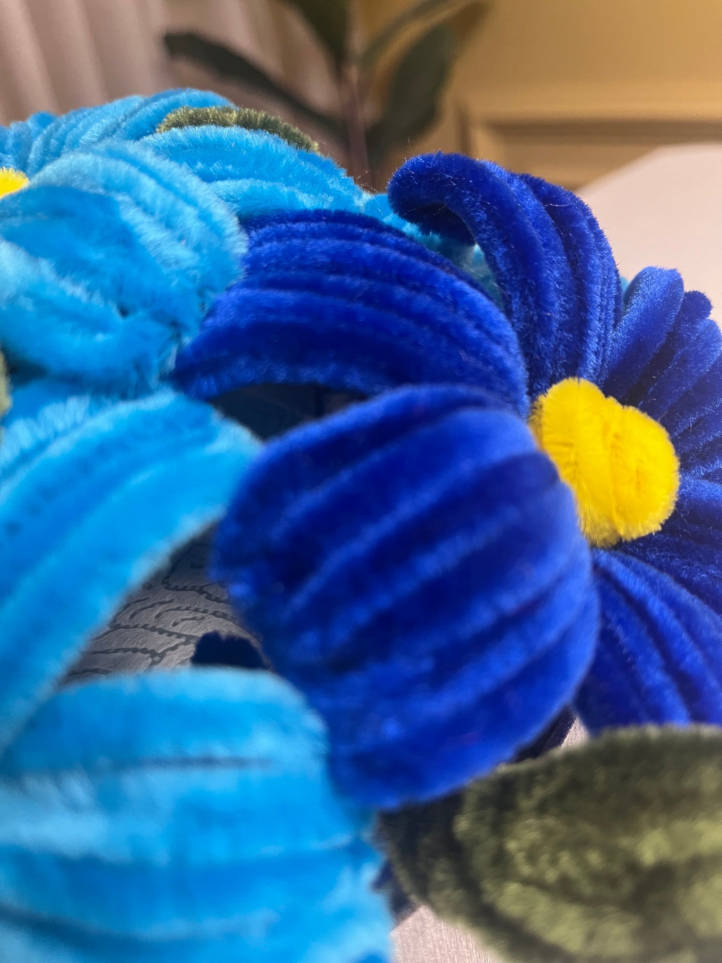 Makro fotografija plavih žičanih cvetova sa žutim središtima i zelenim listovima, prikazuje detaljnu teksturu fuzzy žice i ručnu izradu svakog cveta