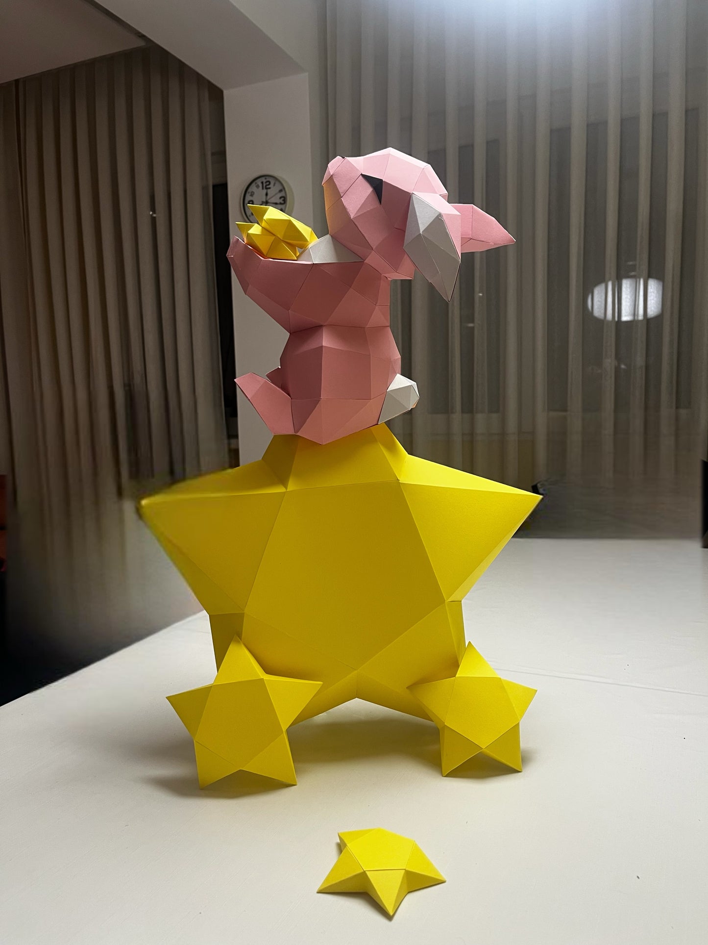 3D papercraft skulptura zečića koji sedi na žutoj zvezdi – roze i žuta paleta, prikaz na stolu