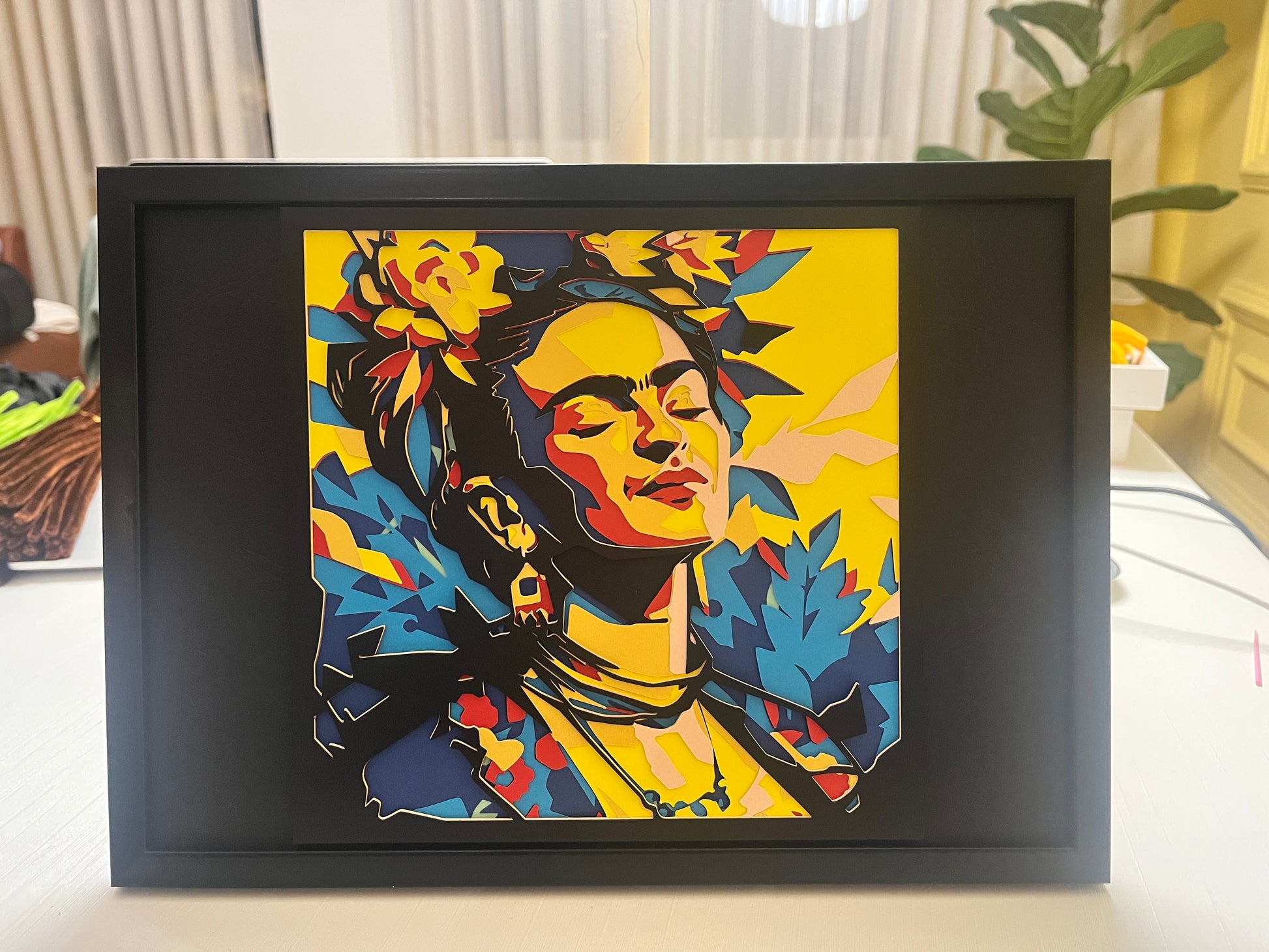 Frontalni prikaz višeslojnog papercut portreta Frida Kahlo u pop-art stilu, crni okvir, žuta pozadina, cveće u kosi