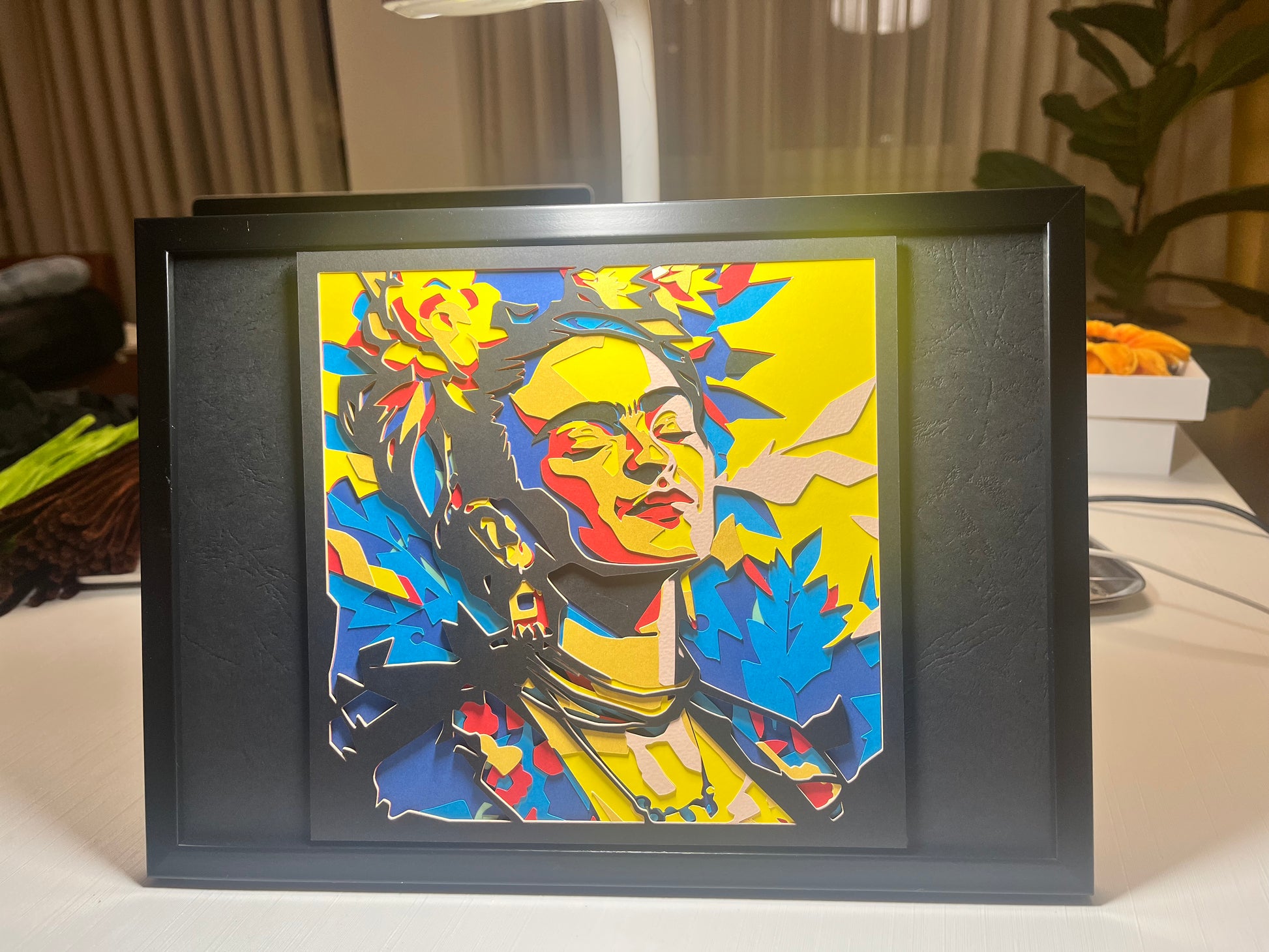 Višeslojna papercut dekoracija Frida Kahlo u crnom okviru, kosi ugao sa lampom u pozadini, žuta pozadina sa plavim i crvenim detaljima