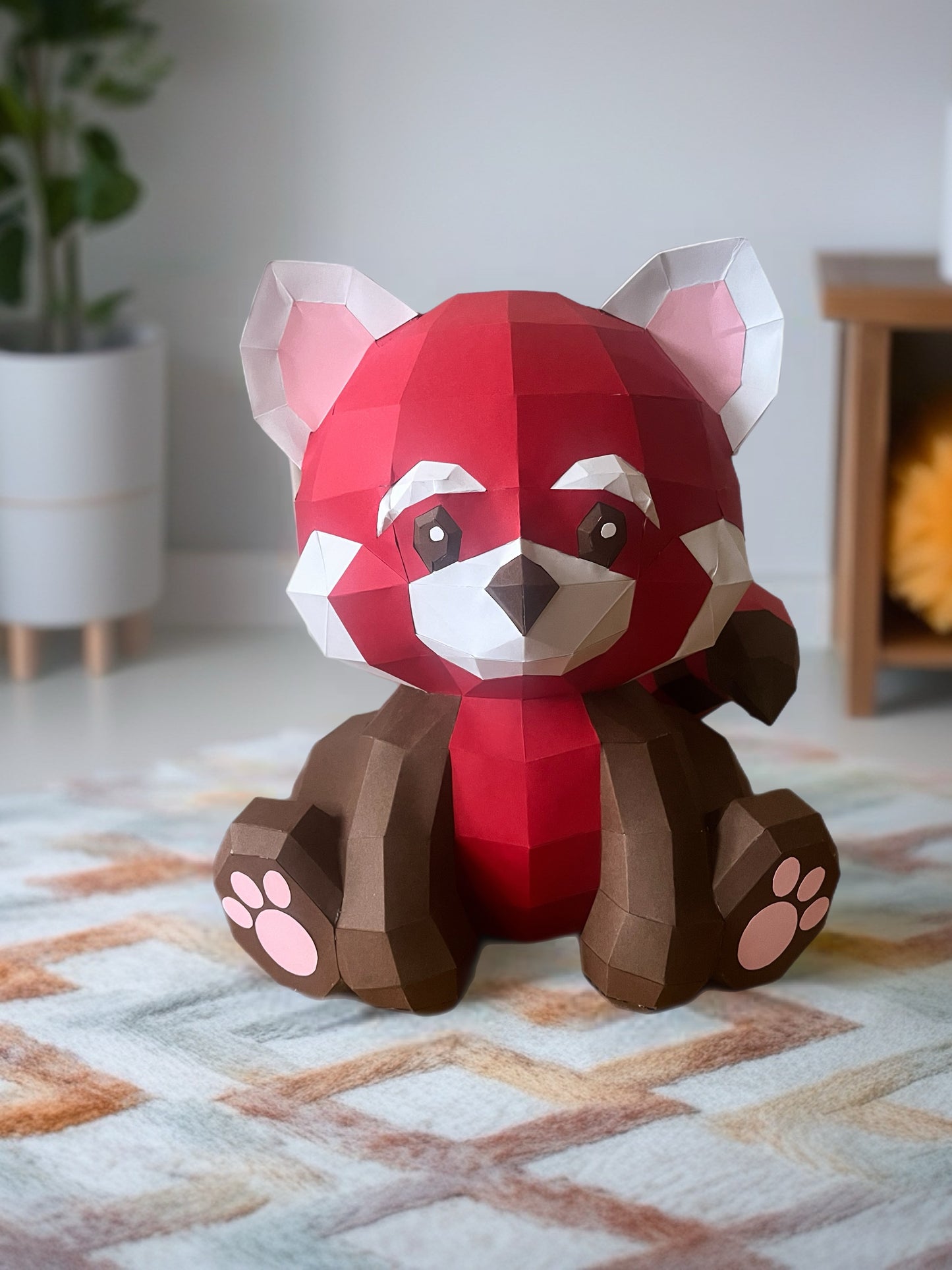 Low poly 3D papercraft skulptura crvene pande koja sedi, crvena i braon boja sa roze šapicama, lifestyle prikaz na šarenom tepihu