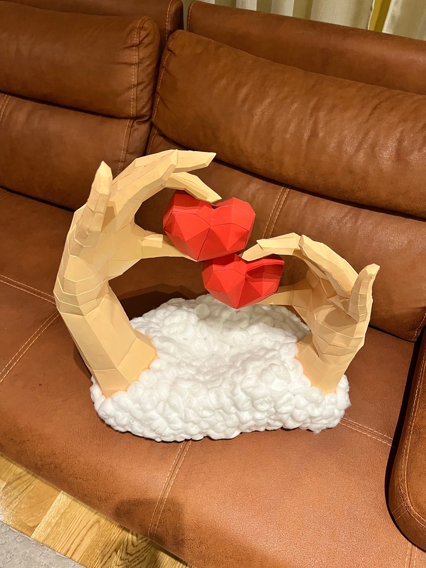 Frontalni prikaz low poly 3D skulpture dve ruke sa dva crvena srca na kožnoj sofi, bela osnova od vate