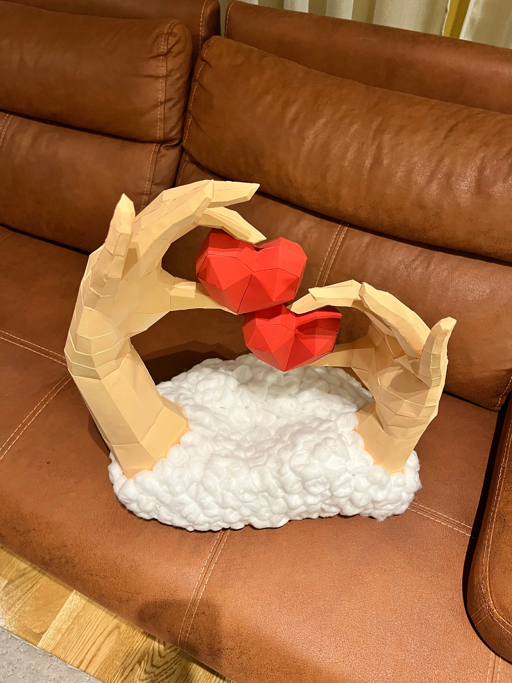 Frontalni prikaz low poly 3D skulpture dve ruke sa dva crvena srca na kožnoj sofi, bela osnova od vate