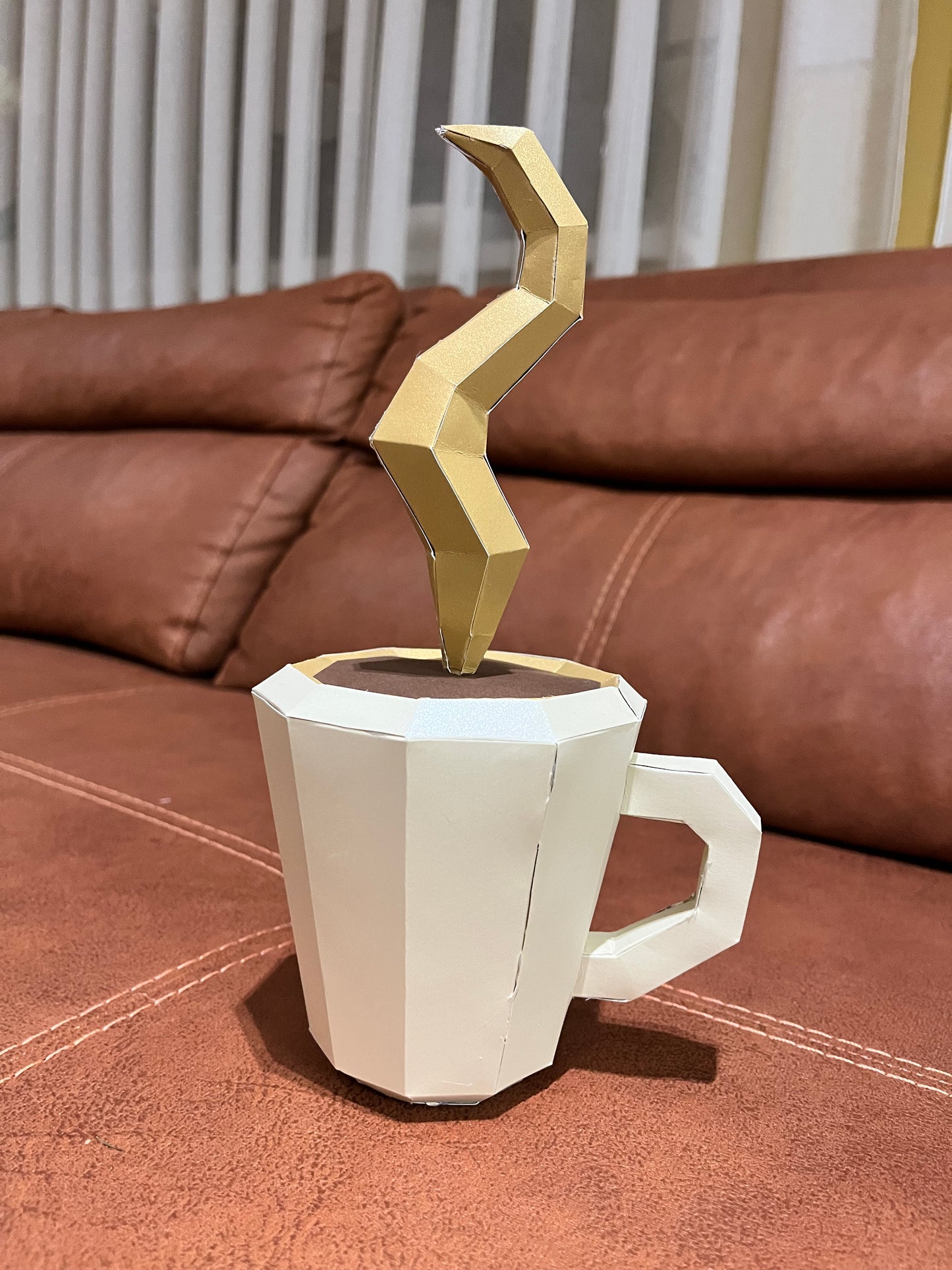 Frontalni krupniji prikaz low poly papercraft šolje kafe sa zlatnom parom koja se vijuga, bela šolja na kožnoj sofi
